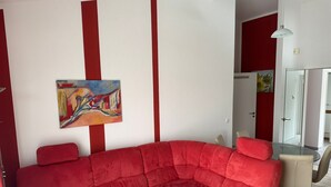 Een rode modulaire bank in een moderne woonkamer met een glazen salontafel, een schilderij aan de muur en een eethoek met een stoel en tafel.