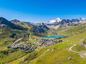 A mountainous landscape with a town nestled in a valley, featuring a lake and snow-capped peaks in the background.
