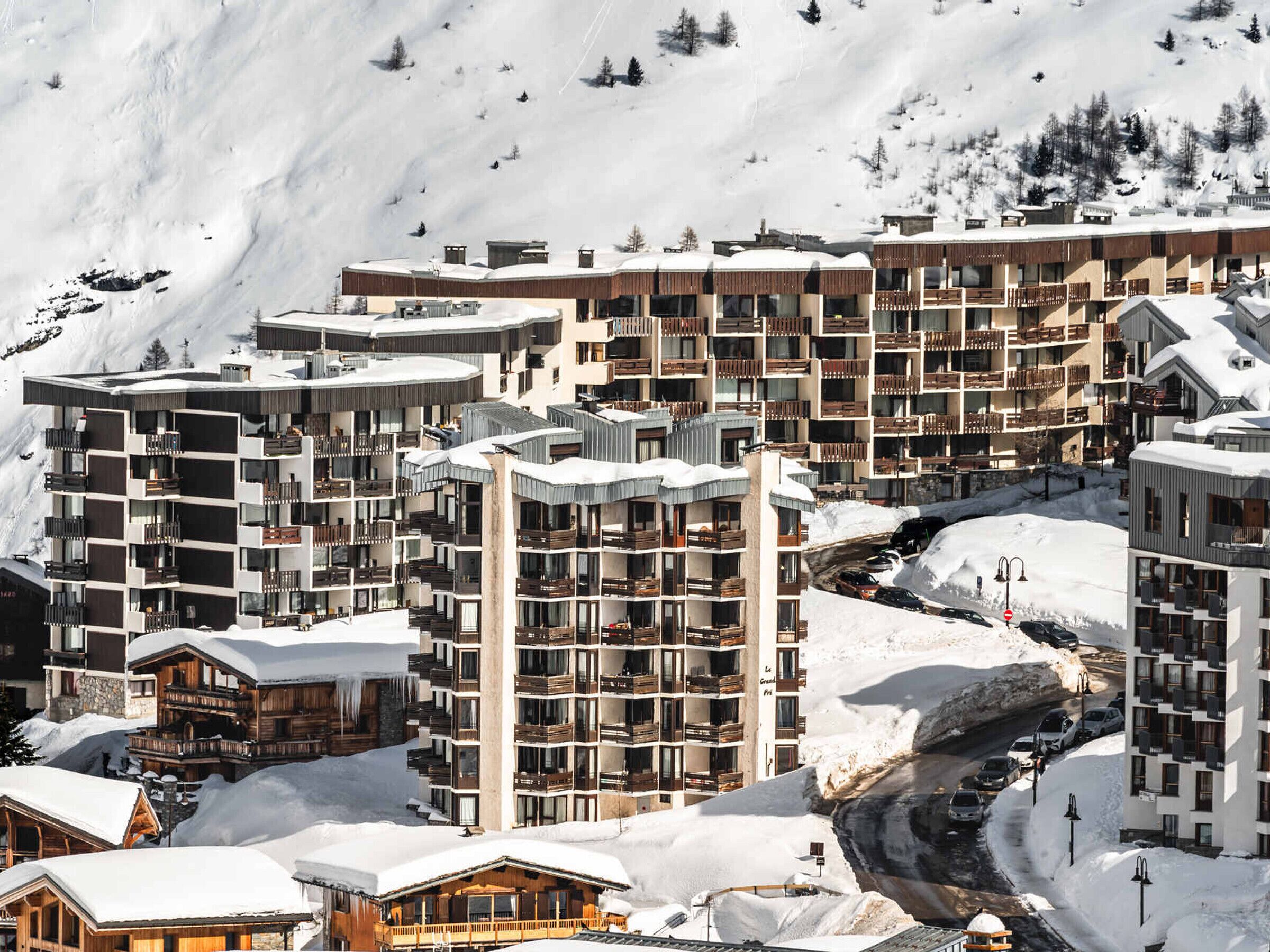 Ein verschneites Bergresort mit mehrstöckigen Gebäuden, darunter ein Hotel mit Balkonen und eine Seilbahn im Hintergrund.