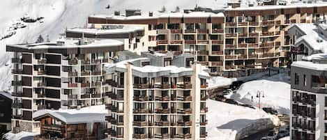 A snow-covered mountain resort with multi-story buildings, including a hotel with balconies and a ski lift visible in the background.