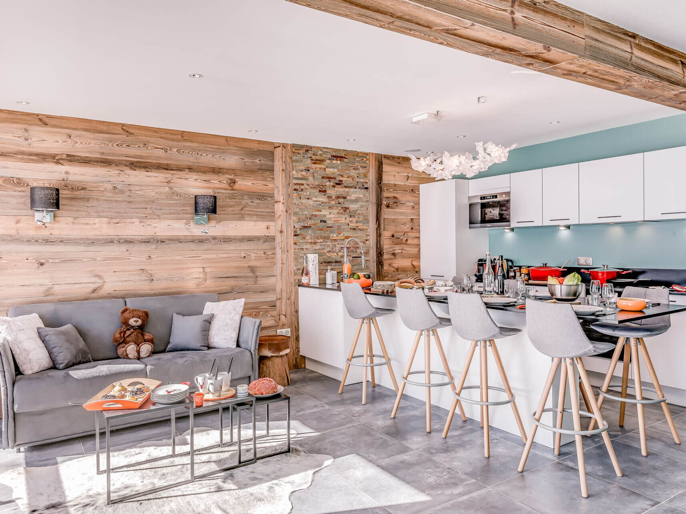 A modern living room with a grey sofa, a wooden accent wall, and a kitchen area with white cabinets and a blue countertop.
