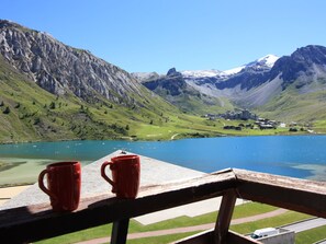 Un lac avec des montagnes en arrière-plan, une terrasse en bois avec deux tasses rouges et un ciel dégagé.
