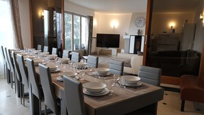 Un comedor moderno con una larga mesa dispuesta para seis personas, rodeada de sillas. Se ve una cocina con televisor y reloj al fondo.