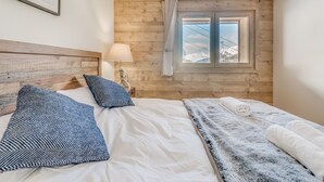 Ein Schlafzimmer mit einem Holzkopfende, einem Bett mit weißer Bettwäsche, einer gemusterten Tagesdecke und einem Fenster mit Blick auf schneebedeckte Berge.