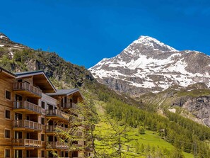 Ein Holzgebäude mit Balkonen vor einer verschneiten Berglandschaft und blauem Himmel.
