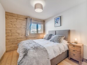 A bedroom with a wooden wall, a bed with a white duvet and grey pillows, a bedside table with a lamp, and a window with a view of snow-capped mountains.
