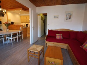 A cozy living room with a red sofa, a wooden coffee table, and a small stool. A dining table with chairs is in the background, and a kitchen is visible through an open doorway.