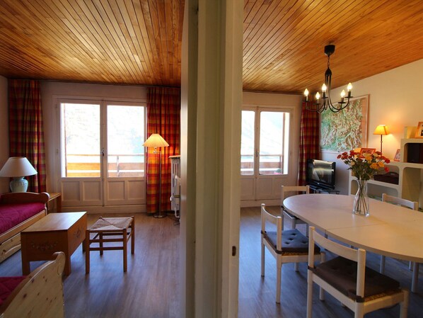 A cozy room with wooden flooring, a dining table with chairs, a sofa, a TV, and a window with curtains.