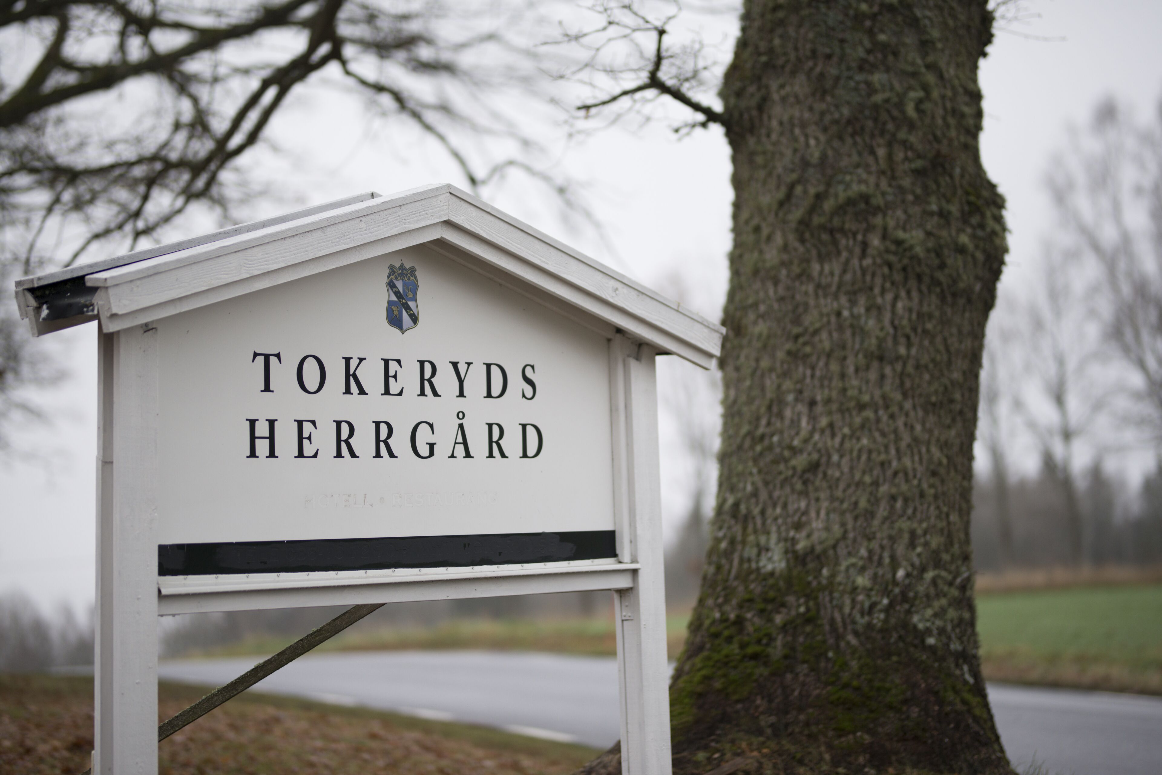 Un letrero blanco con la inscripción 'TOKERYDS HERRGÅRD', sujeto a un marco de metal con techo, al lado de un árbol.