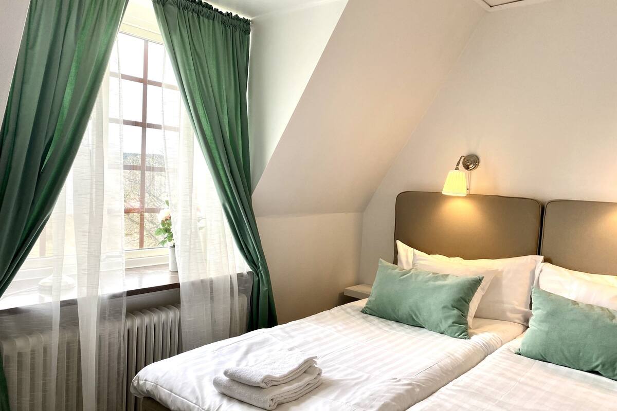 Hotellihuone, jossa on sänky, tyynyjä, yöpöytä, lamppu ja ikkuna, jossa on verhot.