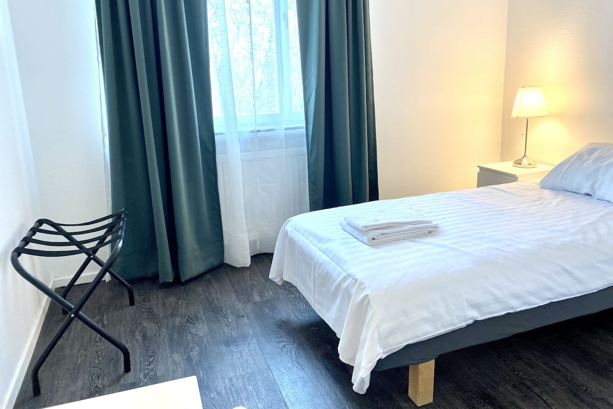 Hotellihuone, jossa on sänky, tuoli, yöpöytä lampun kanssa ja ikkuna, jossa on verhot.