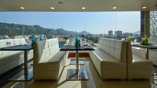 A modern dining area with a city view, featuring a long table and two white cushioned seating areas. - Hotel Atlantica Ajmer (Ajmer)