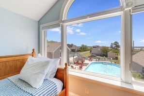 A bedroom with a large window, a bed with white pillows, and a view of a swimming pool and neighborhood.