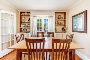 A dining room with a wooden table and chairs, a window with curtains, and shelves with decorative items.