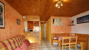 A cozy room with a wooden ceiling, a checkered floor, a sofa with a plaid blanket, a wooden table with chairs, and a kitchen area in the background.