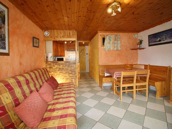 Ein gemütliches Zimmer mit Holzboden, einem Schachbrettmuster-Bodenbelag, einem Sofa mit kariertem Überwurf, einem Holztisch mit Stühlen und einer Küchenzeile im Hintergrund.