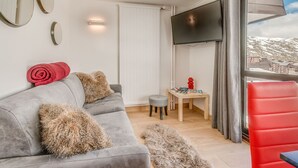 Ein modernes Wohnzimmer mit einem grauen Sofa, flauschigen Kissen, einem roten Sessel, einem kleinen Holztisch mit Büchern und einem an der Wand befestigten Fernseher.
