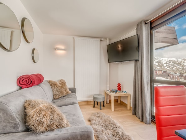 Una sala de estar moderna con un sofá gris, almohadas mullidas, un sillón rojo, una pequeña mesa de madera con libros y un televisor montado en la pared.