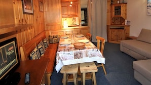 A cozy cabin-style room with wooden walls and ceilings, a dining table set for four, a sofa, and a television displaying a news channel. - Large studio for 4 ppl. with balcony and lake view in Tignes (Tignes)