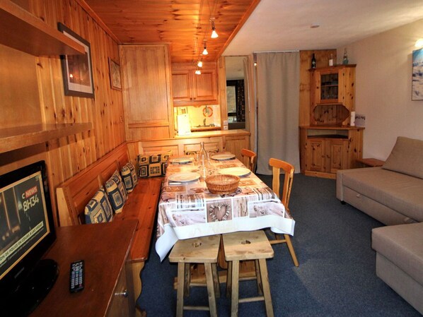 Une pièce confortable, aménagée comme un chalet, avec des murs et un plafond en bois, une table à manger pour quatre personnes, un canapé et un téléviseur diffusant une chaîne d’information.