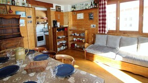A cozy cabin-style room with wooden walls, a dining table set with plates and glasses, a sofa, and a kitchen area with a stove and shelves of bottles. - Traditional Chalet in Tignes - 3 rooms, 6 people, WiFi included (Tignes)