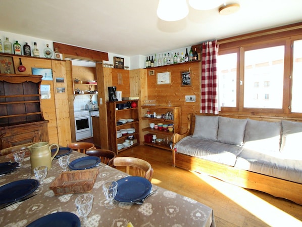 A cozy cabin-style room with wooden walls, a dining table set with plates and glasses, a sofa, and a kitchen area with a stove and shelves of bottles.
