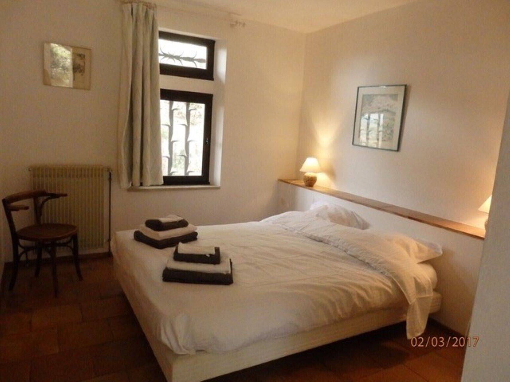 A neatly made bed with folded towels, a chair, a radiator, and a window with curtains.