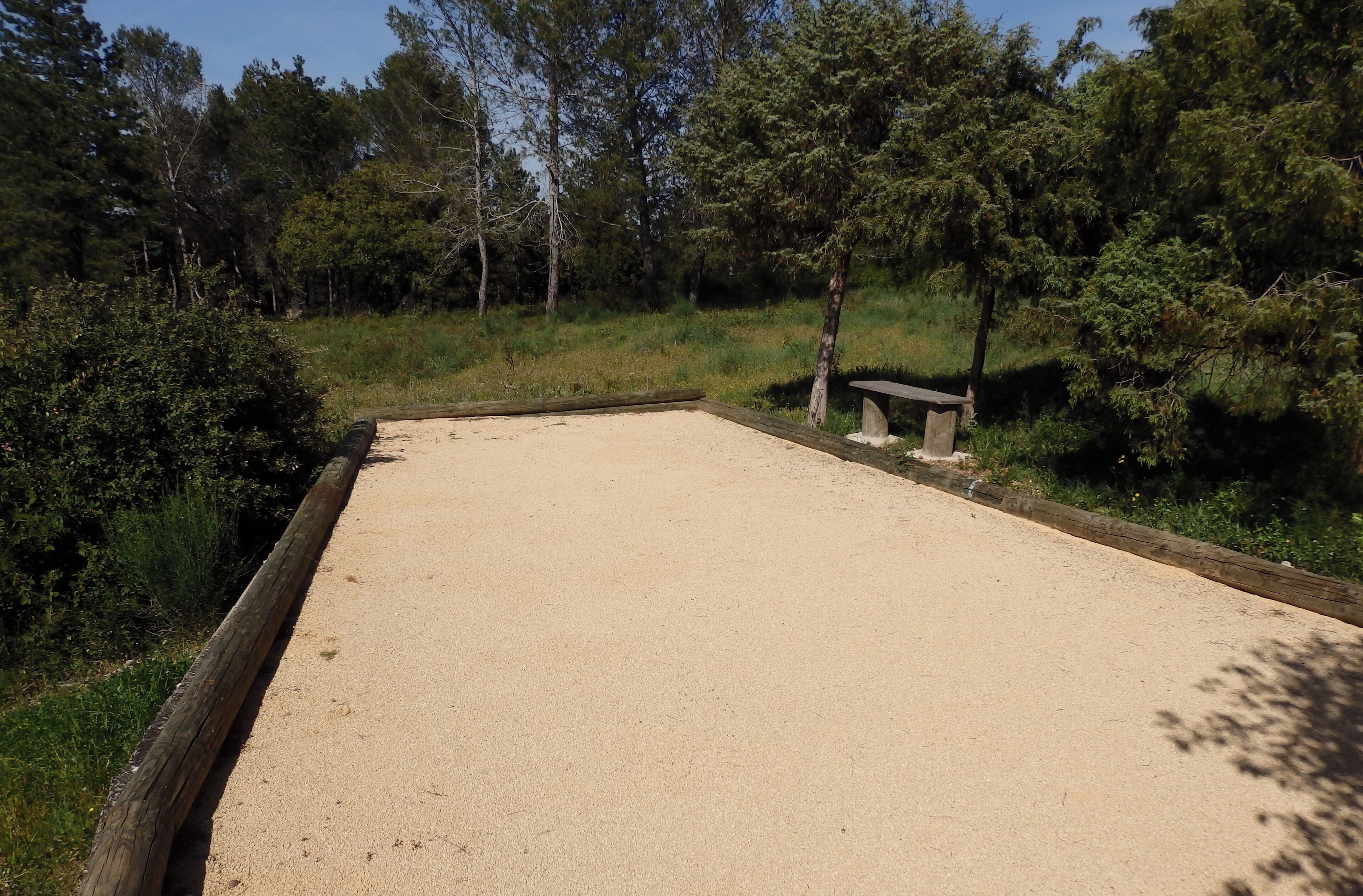 Un espace sablonneux avec un banc en bois, entouré d’une végétation luxuriante et d’arbres.