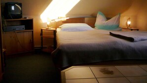 Una habitación de hotel con cama, televisor en un estante, mesita de noche con lámpara y suelo de baldosas.