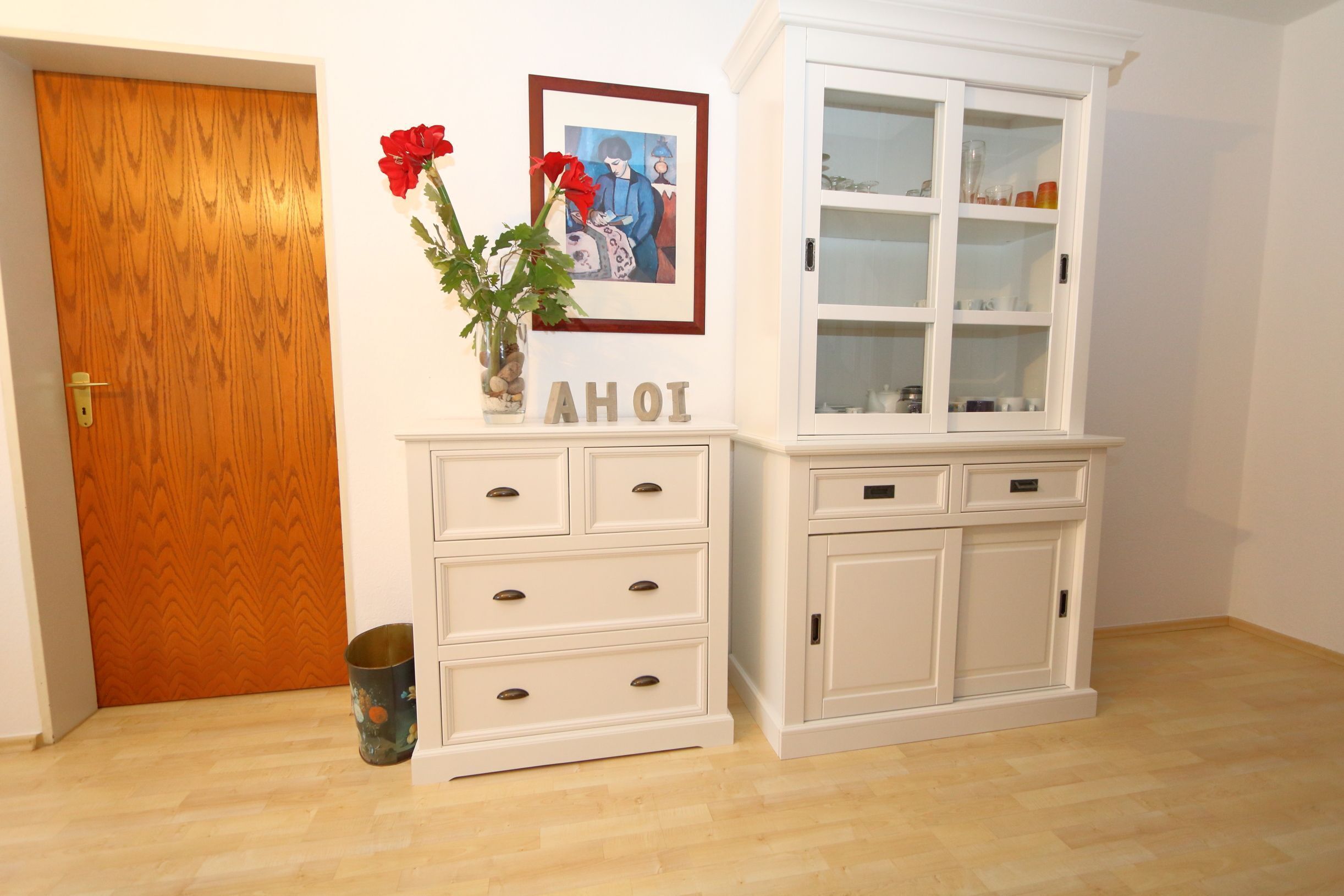 Una habitación con puerta de madera, un aparador blanco con cajones, un mueble aparador con puertas de cristal y un cuadro con flores rojas.