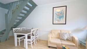A living room with a staircase, a dining table with chairs, a sofa, and a framed picture on the wall. - App Kapitänsstube, Germany (Sylt)