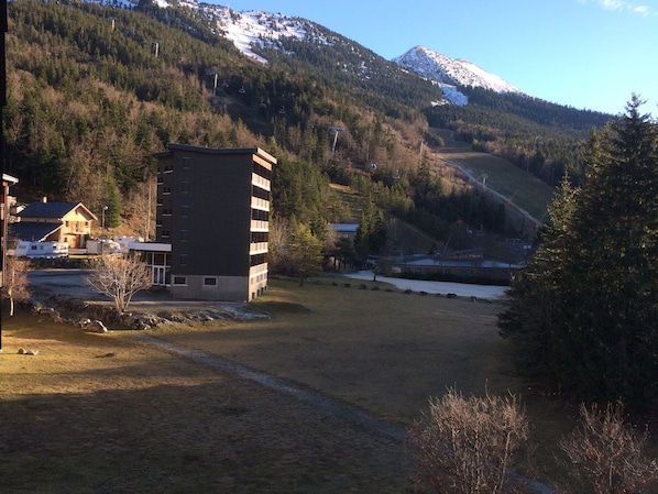 A modern building with a flat roof and multiple windows, surrounded by a grassy area and a forested mountain backdrop.