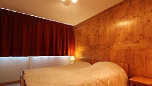 A bedroom with a wooden headboard, a bed with a beige duvet, a nightstand with a lamp, and a window with red curtains. - Spacious 3-room family apt - 2 bathrooms - Lake view balcony (Tignes)