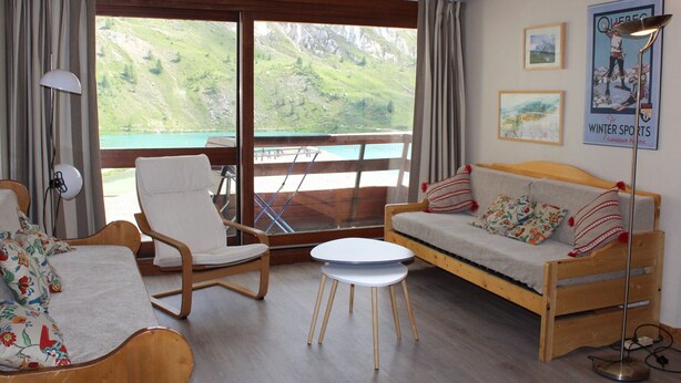 A cozy room with a wooden bed, a white sofa, a wooden chair, and a small table. There is a large window with a view of green hills and a lake. - Spacious 3-room family apt - 2 bathrooms - Lake view balcony (Tignes)