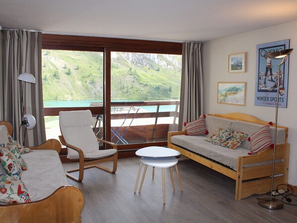 Ein gemütliches Zimmer mit einem hölzernen Bett, einem weißen Sofa, einem Holzstuhl und einem kleinen Tisch. Es gibt ein großes Fenster mit Blick auf grüne Hügel und einen See.