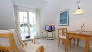 A room with a dining table, chairs, a television, and a window with curtains. - Spieker Wenningstedt (Wenningstedt-Braderup (Sylt))