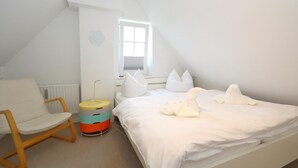 A bedroom with a sloped ceiling, a bed with white bedding, a chair, a bedside table, and a window. - Spieker Wenningstedt (Wenningstedt-Braderup (Sylt))