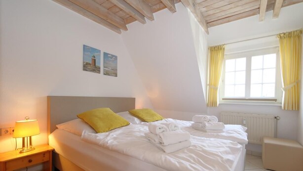 A neatly made bed with white linens and two yellow pillows, a bedside table with a lamp, and a window with yellow curtains. - Syltnest Munkmarsch, Germany (Sylt-Ost)