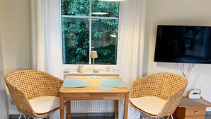 A room with two wicker chairs, a wooden table, a TV on the wall, and a small radio on a side table.