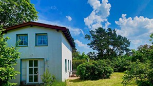 A two-story house with a red roof and white walls, surrounded by lush greenery and trees.
