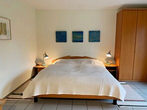 A bedroom with a bed, two bedside tables with lamps, a wooden wardrobe, and three wall-mounted paintings.
