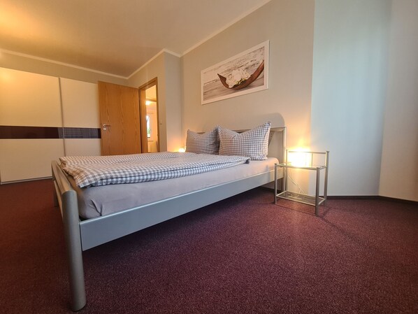 A single bed with a checkered bedspread, a bedside table with a lamp, a wooden wardrobe, and a framed picture on the wall.