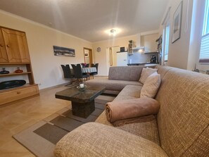 A living room with a sofa, a coffee table, a dining table with chairs, a kitchen area, and a clock on the wall.