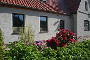 A building with a red-tiled roof, two windows with shutters, and a garden with various plants and flowers.