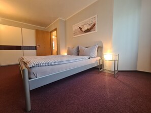 Uma cama de solteiro com colcha xadrez, mesa de cabeceira com luminária, guarda-roupa de madeira e quadro na parede.