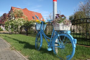 Ein blaues Fahrrad mit Korb und Blumentopf, abgestellt auf einer Rasenfläche mit einem Pflasterweg und einem Metallzaun im Hintergrund.