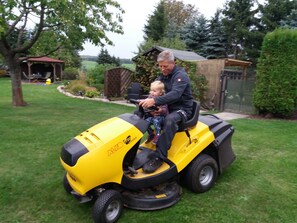 Ein gelber Rasenmäher mit einem Kind und einem Erwachsenen darauf, umgeben von einem gepflegten Rasen und einem Garten.