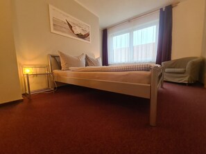 Ein Einzelbett mit Metallgestell, ein Nachttisch mit Lampe, ein Sessel und ein Fenster mit Vorhängen.