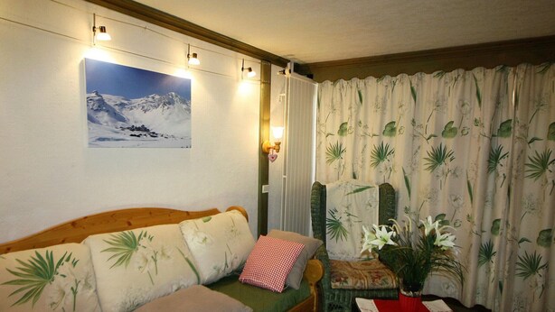 A cozy living room with a wooden sofa, a green cushion, and a floral-patterned armchair. A framed picture hangs on the wall, and a window with curtains is visible. - Classic studio for 4, south terrace lake view (Tignes)