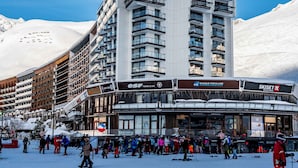 A multi-story hotel with balconies, a ski resort in the background, and a crowd of people in winter attire. - Classic studio for 4, south terrace lake view (Tignes)